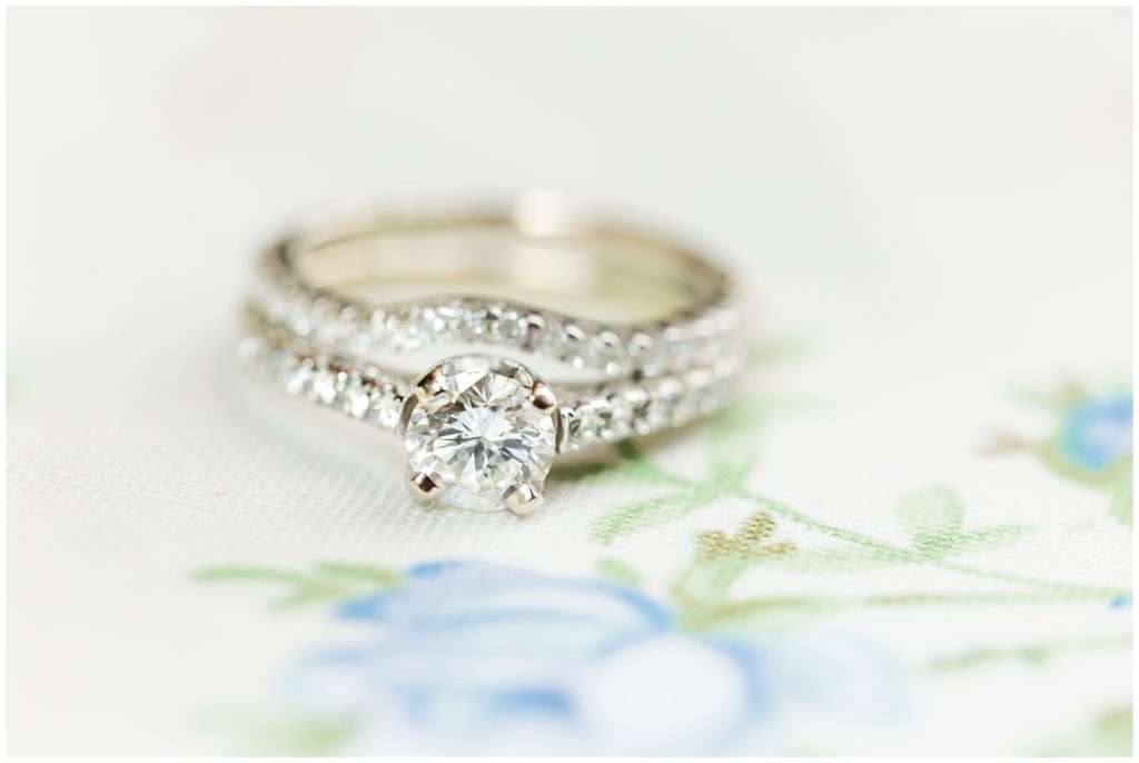 The bride's diamond engagement and wedding band is displayed with a distant background of white and light blue florals. 