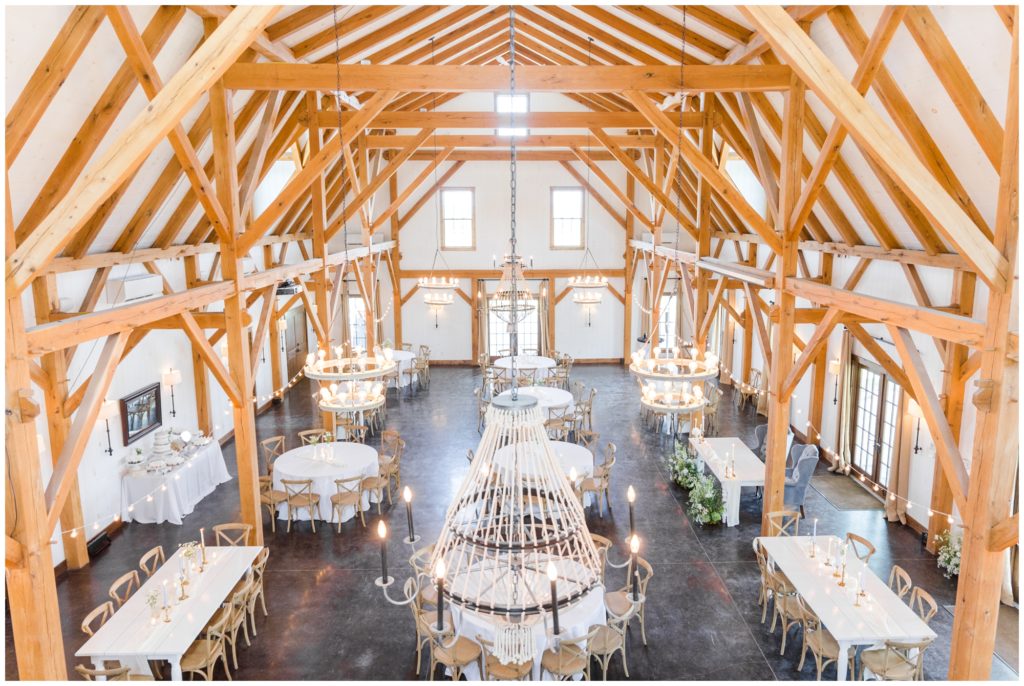 The interior of Blue Bell Farm is shown set up for the reception - white tables with simple floral arrangements surrounded by wooden chairs. 