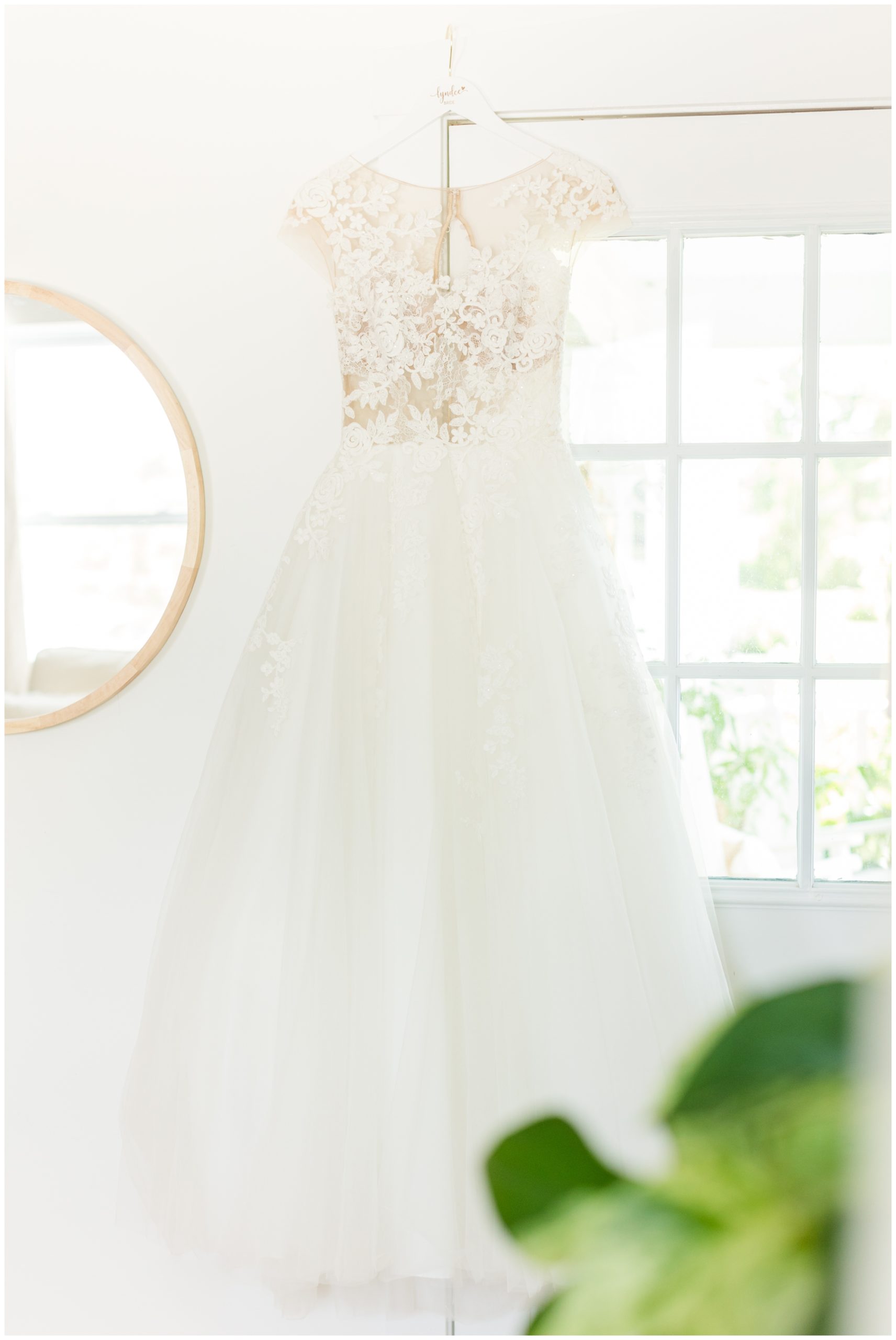 The bride's lace wedding dress is hung on a white custom hanger in front of a window.