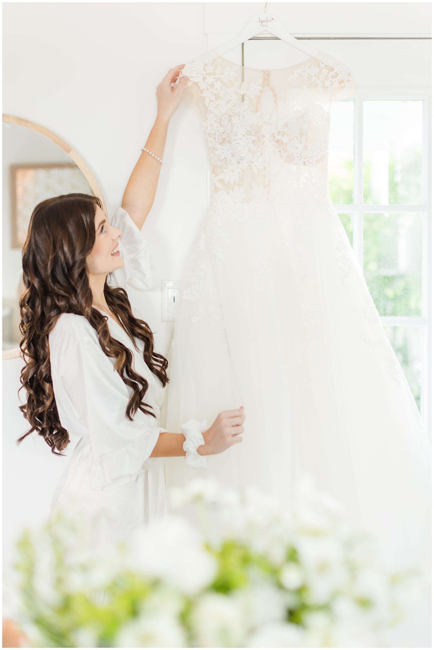 The bride's lace wedding dress is hung on a white custom hanger in front of a window. The bride poses with it, smiling. 