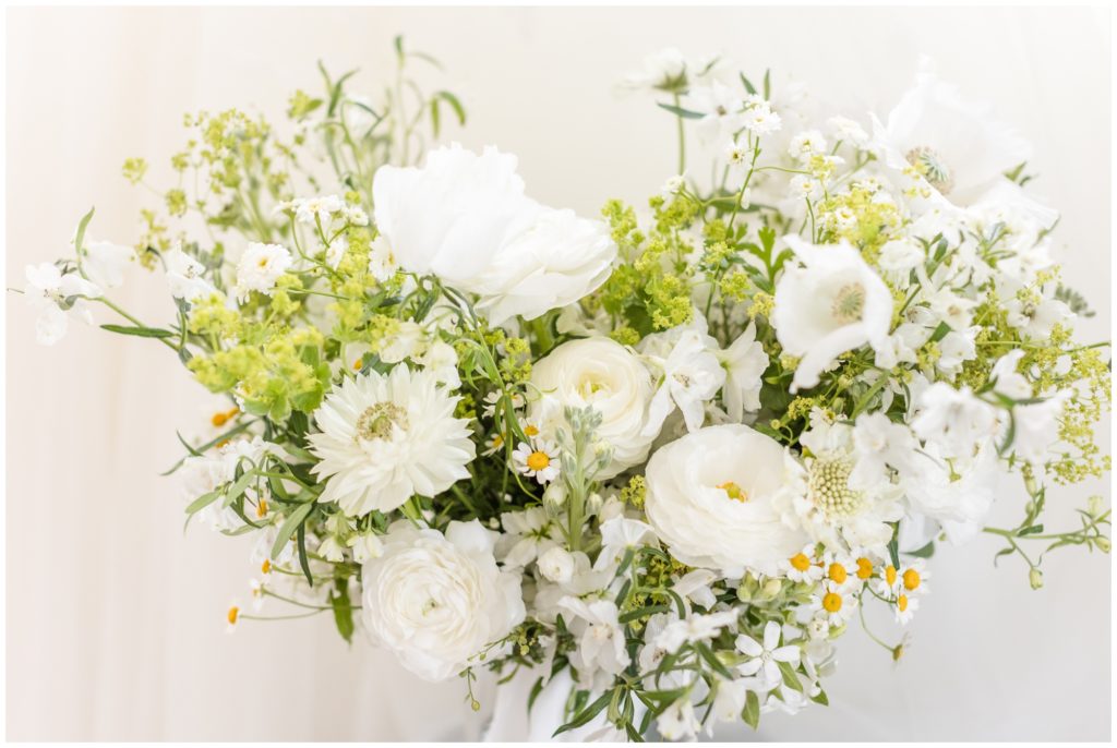 The bride's wedding bouquet is pictured. The flowers are simple and all white, with pops of delicate greenery and some tiny white daisies with yellow centers. 