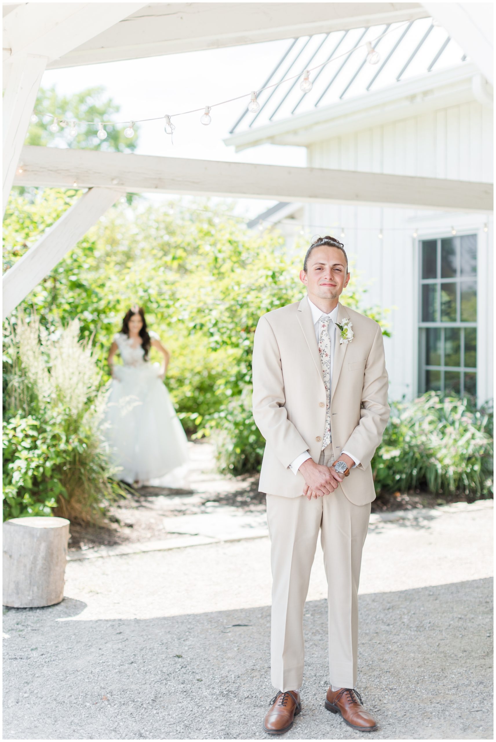 The groom poses in his khaki suit with white shirt and floral tie.  His bride is unfocused in the background as she walks up for the first look. 