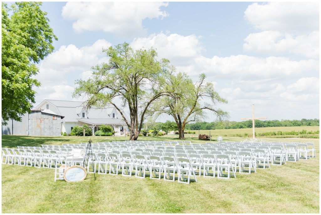 The ceremony site is shown for this Blue Bell Farm wedding, complete with simple wooden cross surrounded by babies breath set up in front of white chairs. 