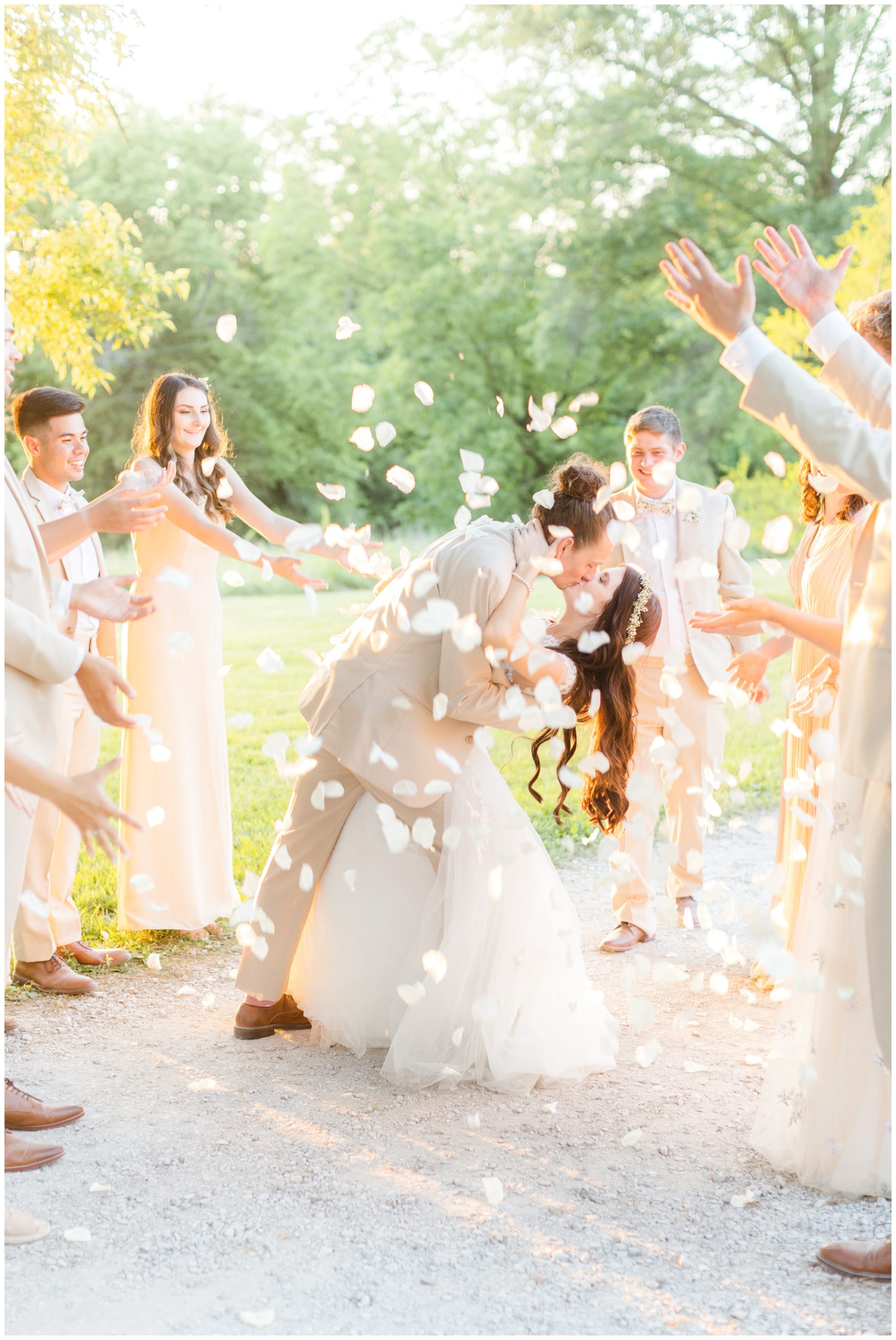 The wedding exit is pictured. The groom is dipping his new bride back with a kiss, and the wedding party is showering them with soft white flower petals. 