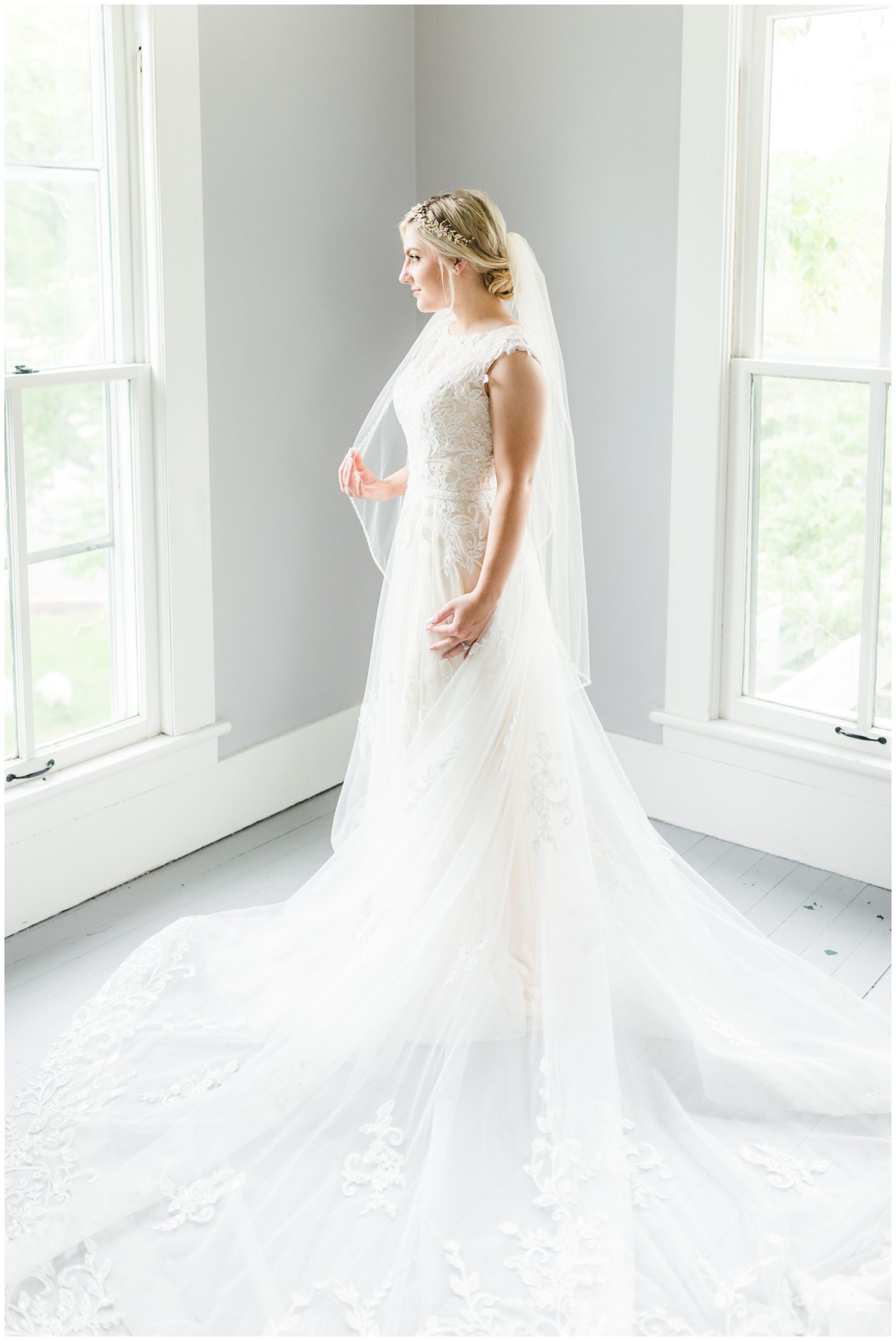 The bride stands in her white lace gown in front of a window. She holds her veil in her fingertips. 