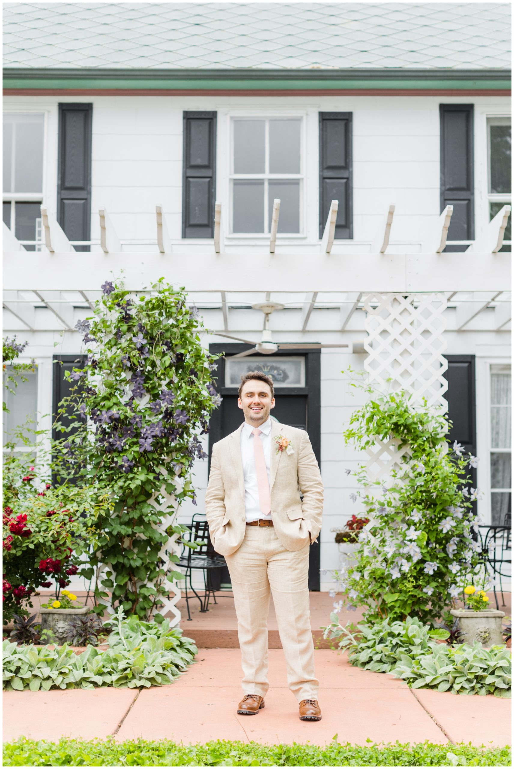 The groom poses in his tan linen suit on an exterior porch at defiance vineyard. He is wearing a pale pink tie with a boutonniere (orange and white flowers plus greenery) tied with a pink ribbon.  