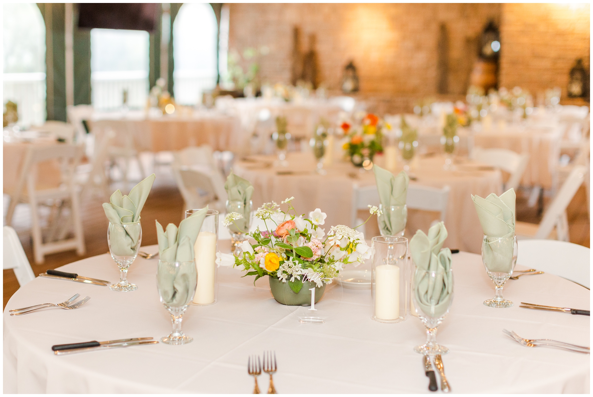 The reception space is pictured. The tables are covered in a pale linen and are surrounded with white wooden chairs. There are short floral arrangements on the tables. 