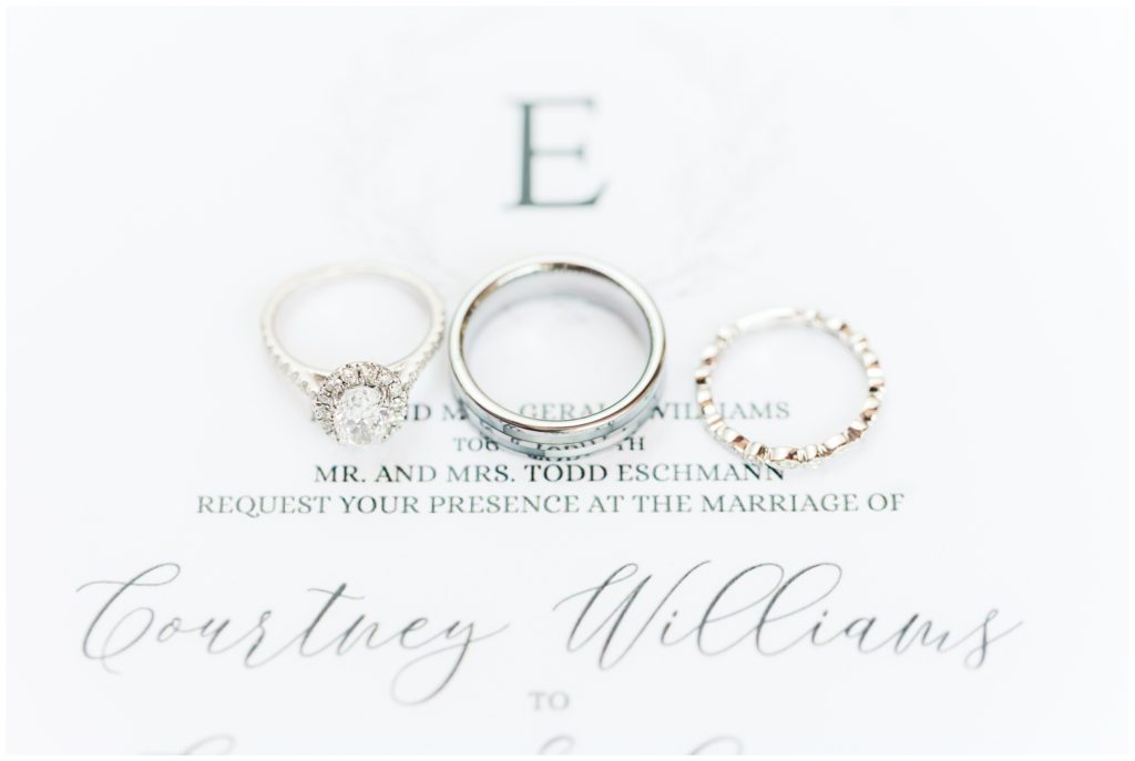 A white gold and diamond engagement ring and wedding band, together with the white gold men's wedding band, are displayed on a the black and white wedding invitation. 