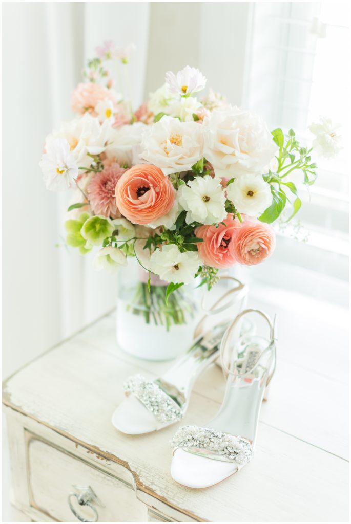 The bride's Badgley Mischka crystal studded wedding shoes are on top of a distressed white dresser, next to a vase of English garden type florals. 