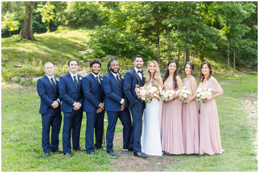 The bridal party is pictured. The bridesmaids are in a blush pink floor-length gown and the men are wearing blue suits. 