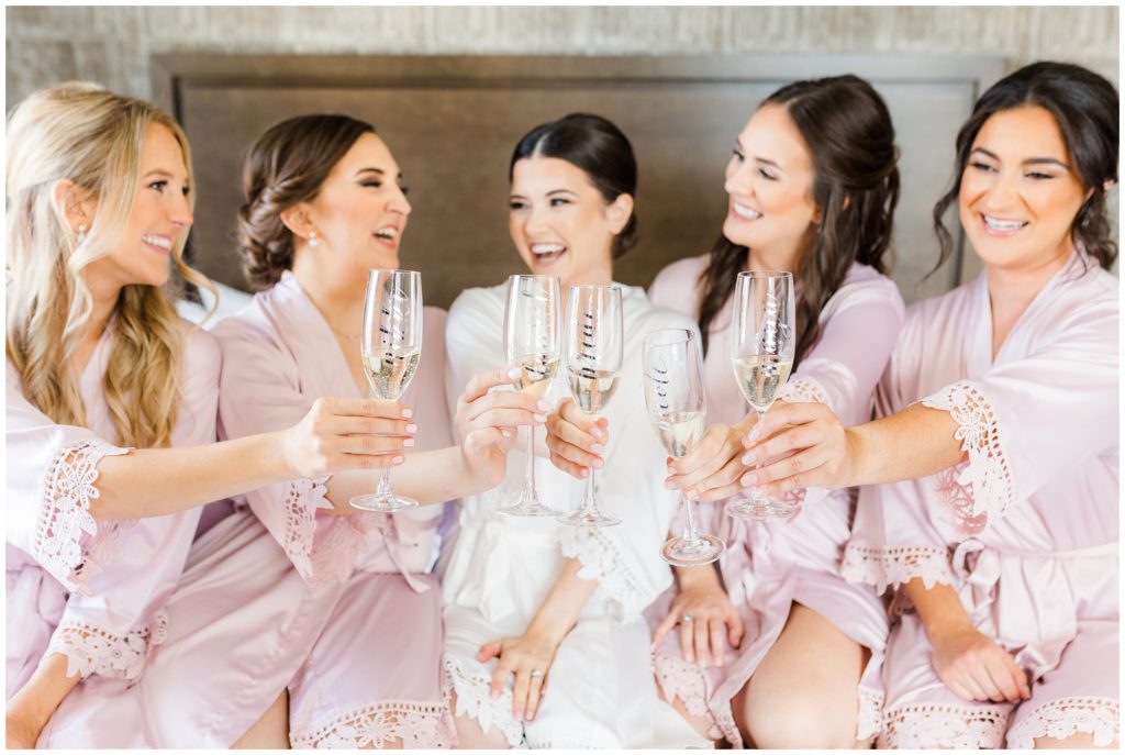 The bride laughs with her attendants in matching robes while they cheers champagne in matching glasses. 